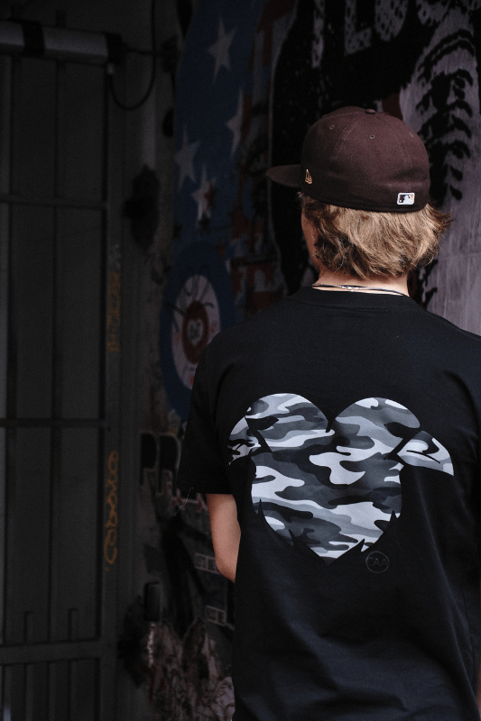 Person wearing a black t-shirt with a camouflage heart design, standing in front of a wall with graffiti.