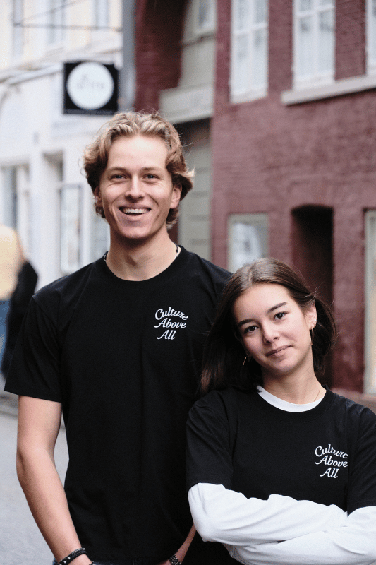 Two people wearing black t-shirts with 'Culture Above All' text standing in front of a building.
