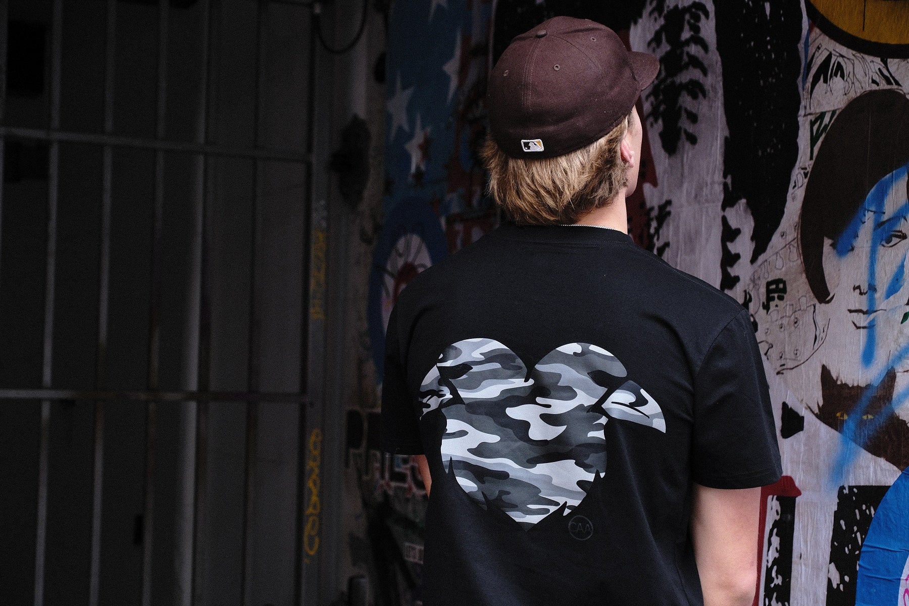 Person wearing a black t-shirt with a camouflage heart design, standing in front of a graffiti wall.
