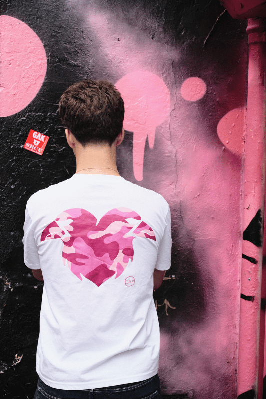 Person wearing a white t-shirt with a pink heart design, standing in front of a graffiti-covered wall.